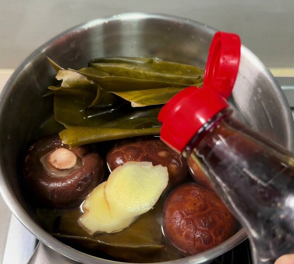 Sauce de poisson versée dune bouteille dans une petite casserole contenant du kombu, des shiitakes et du gingembre émincé.