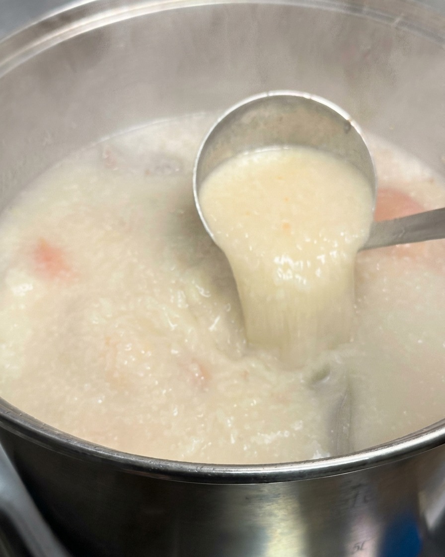 Une louche en métal soulevant une portion de bouillon tonkotsu blanc laiteux et opaque dune grande marmite fumante.
