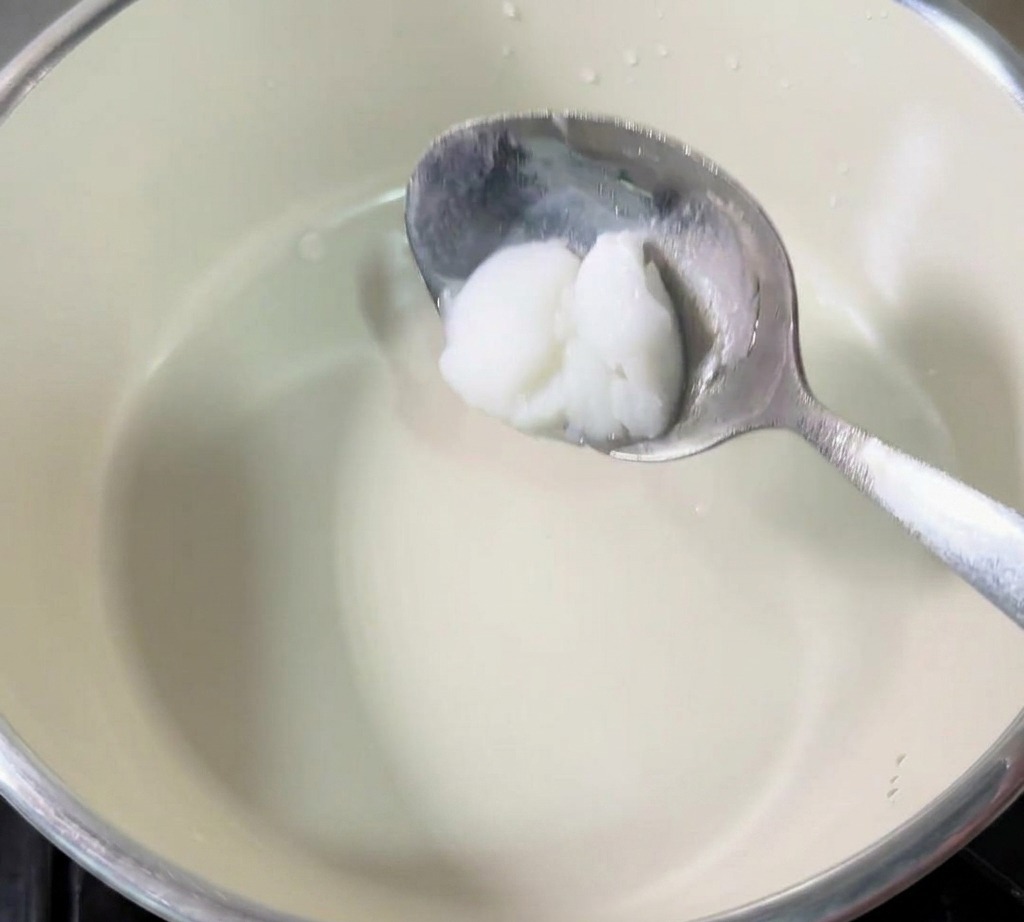 Une cuillère en métal tenant une noix de saindoux blanc et solide au-dessus du centre dune petite casserole de couleur crème.