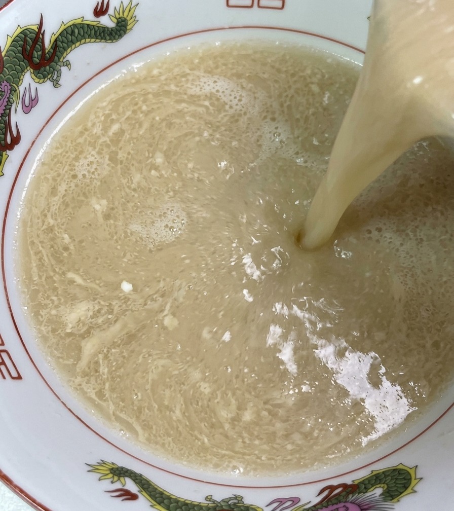 Bouillon dos de porc fumant de couleur ivoire versé dune louche dans un bol de ramen décoré, montrant sa texture riche et soyeuse.
