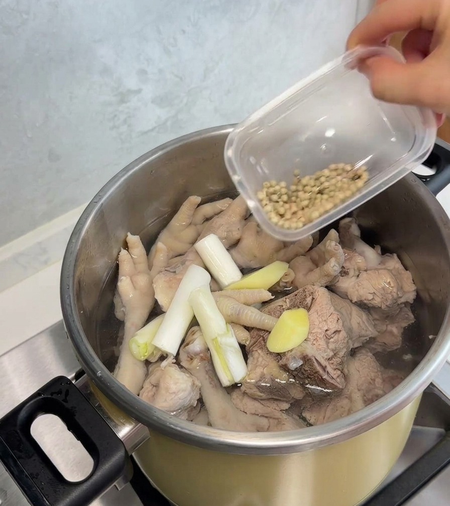 Une main verse des grains de poivre blanc dans une marmite en acier inoxydable contenant des pattes de poulet blanchies, des os de porc, des cébettes et des rondelles de gingembre.