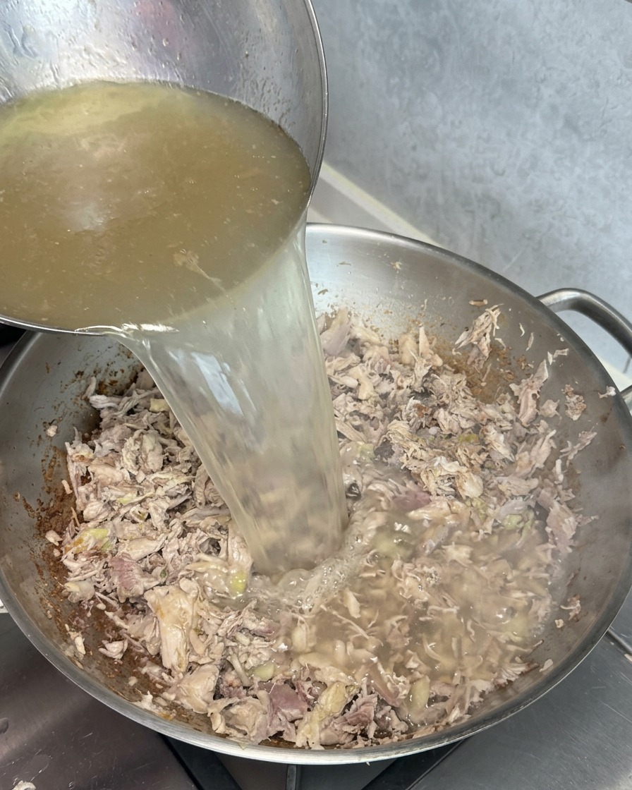 Une louche versant un bouillon doré et clair dans un wok rempli de restes de viande dorée, créant une réaction dun blanc trouble au contact.