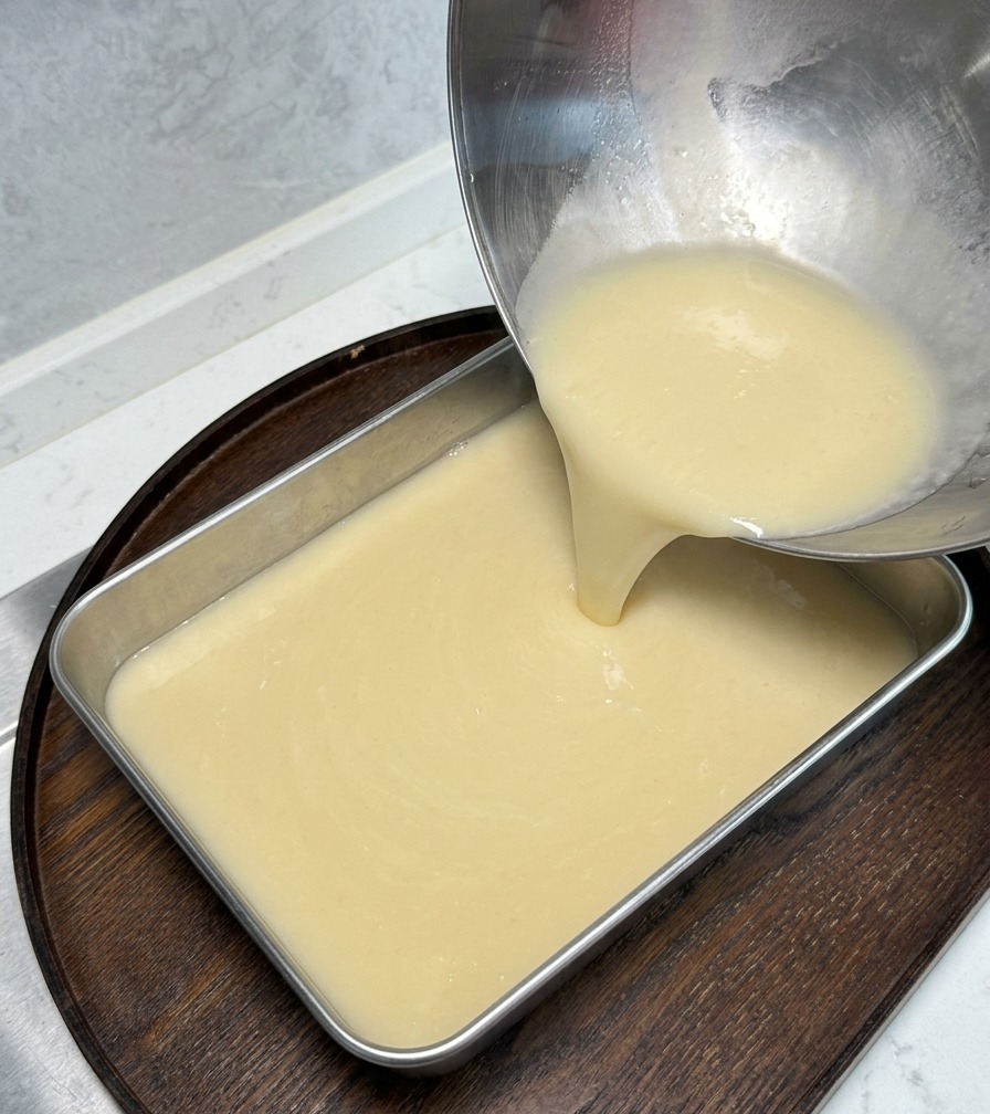 Un bouillon épais de couleur crème est versé dun bol en acier inoxydable dans un plateau rectangulaire en métal posé sur une planche en bois.