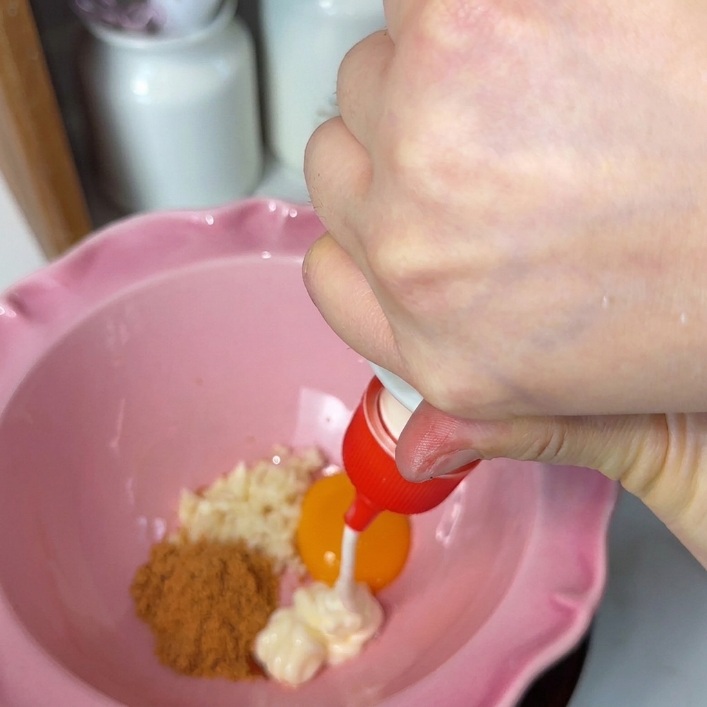 De la mayonnaise blanche est pressée dun embout rouge dans un bol rose cannelé contenant de lail haché, un jaune dœuf orange vif et de la poudre dassaisonnement brune.