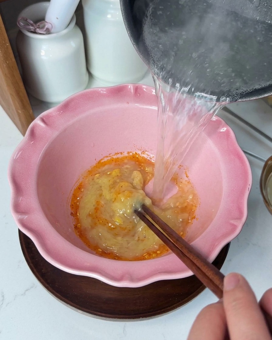 Liquide clair et très chaud versé depuis une casserole dans un bol rose contenant une pâte dassaisonnement jaune crémeuse, remuée avec des baguettes en bois.