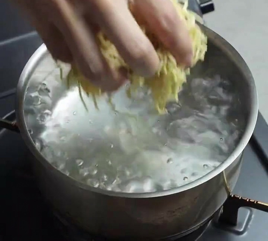 Une main plongeant un nid de nouilles ramen fraîches et jaunes dans une casserole en acier inoxydable remplie deau bouillante.