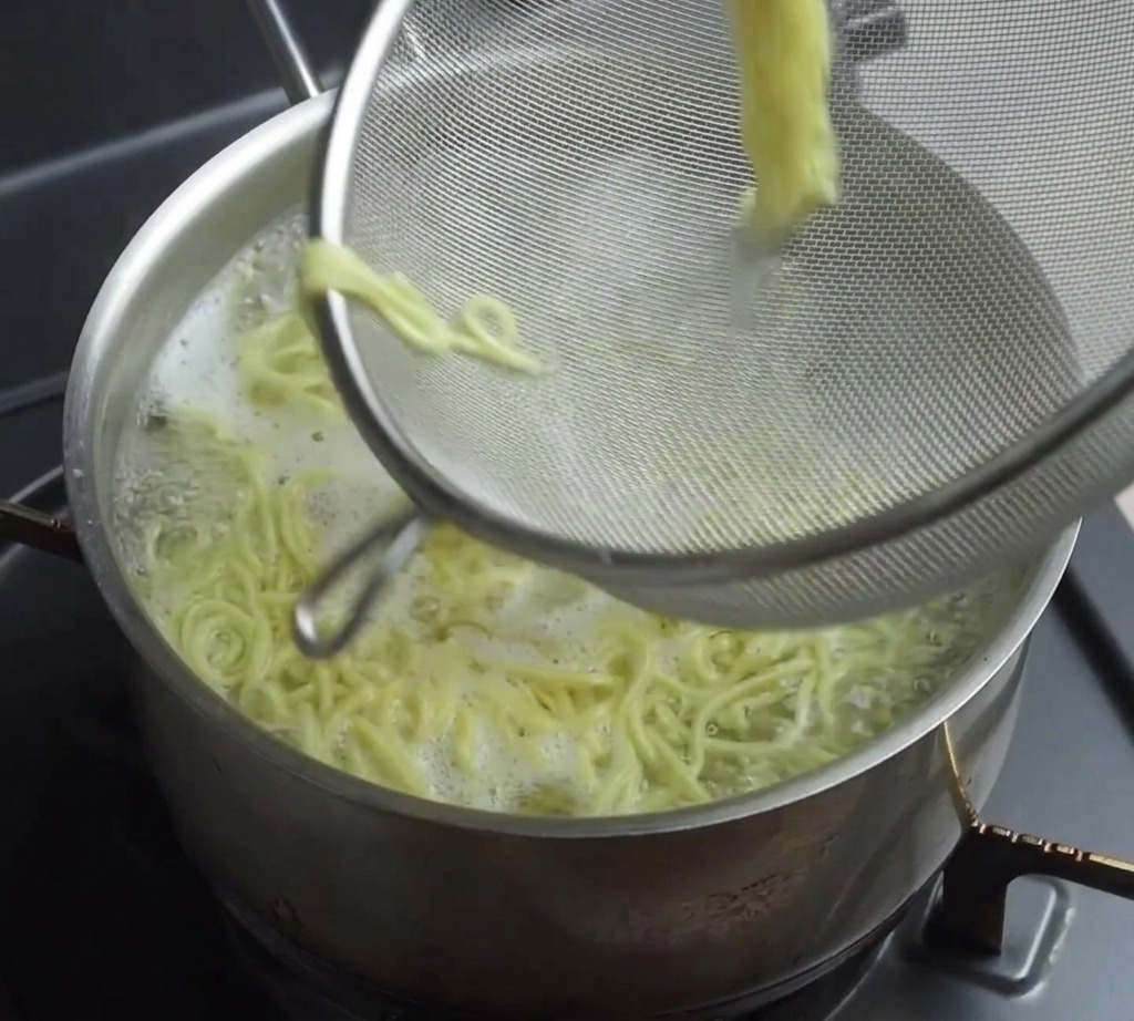 Une passoire à mailles fines soulevant une portion de nouilles ramen jaunes cuites hors dune casserole fumante.