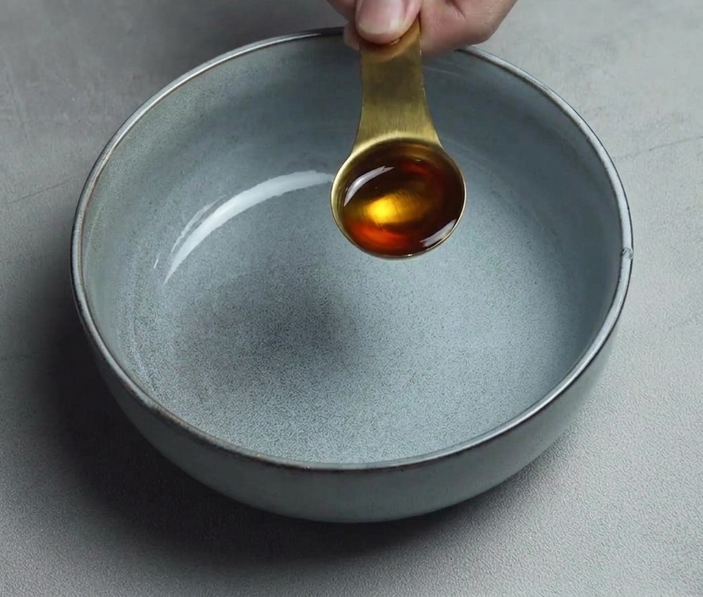Une cuillère à soupe dorée dhuile de sésame versée dans un bol en céramique gris propre et vide.