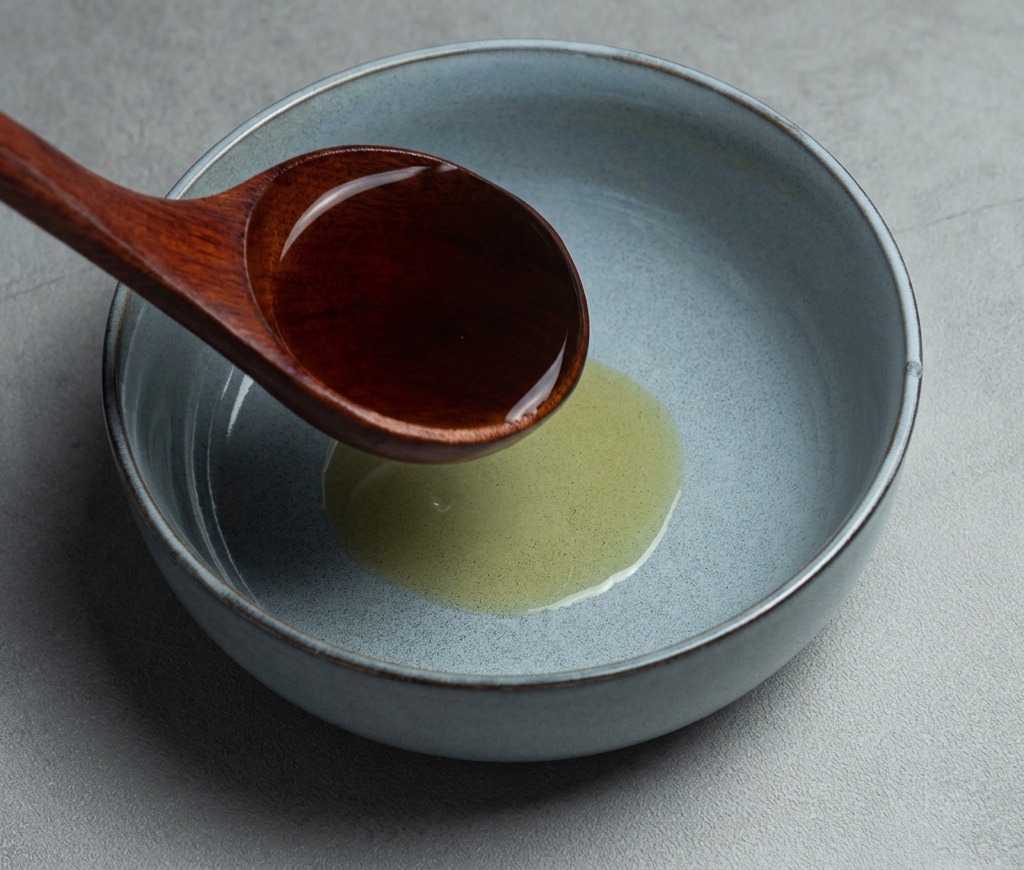 Une louche en bois foncé versant un bouillon de ramen shoyu chaud dans un bol en céramique gris contenant de lhuile de sésame.