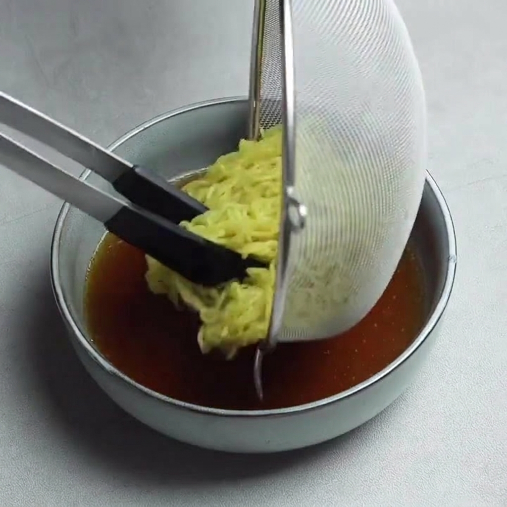Nouilles ramen jaunes transférées dune passoire vers un bol de bouillon foncé savoureux à laide de pinces à poignées noires.