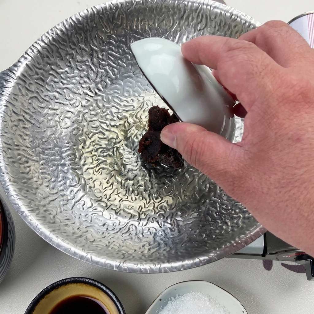 Une main utilisant un petit bol blanc pour déposer une cuillerée épaisse de pâte de miso rouge foncé dans le liquide réduit.