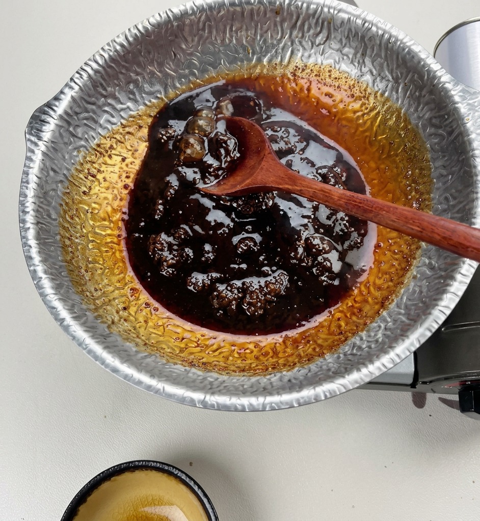 Une cuillère en bois remuant une sauce sombre, épaisse et frémissante dans une casserole en argent martelé.