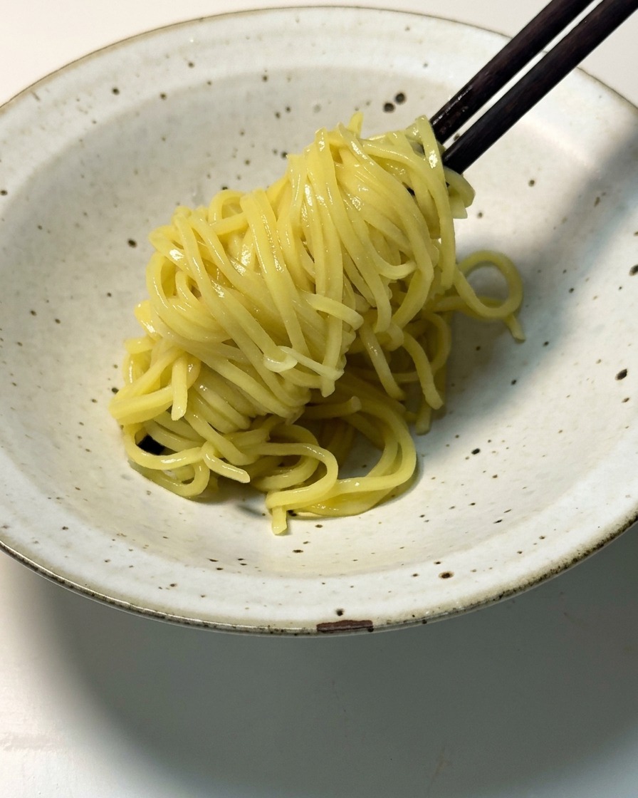 Des nouilles ramen jaunes cuites soulevées avec des baguettes en bois sombre et déposées dans un bol en céramique moucheté.