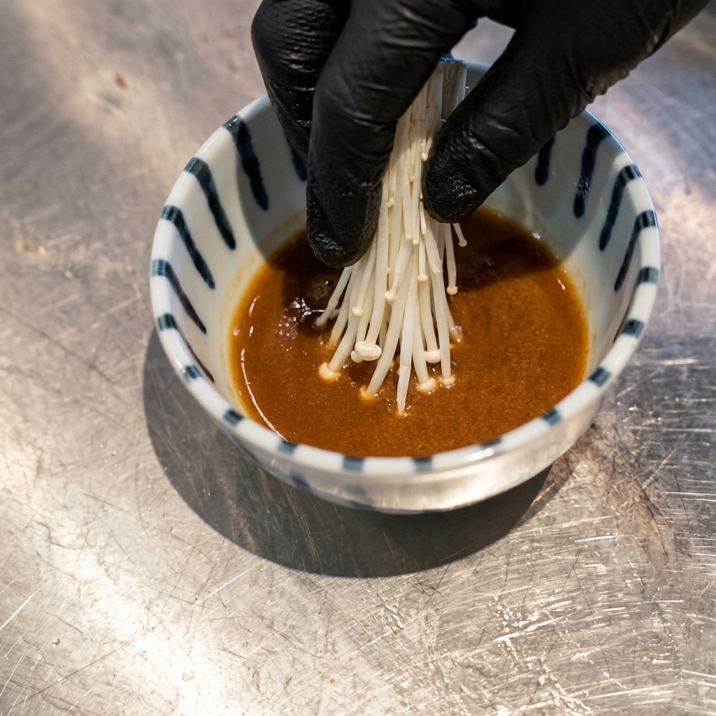 Une main portant un gant noir plaçant un nid de germes de soja blancs dans un bol de sauce de trempage brune.