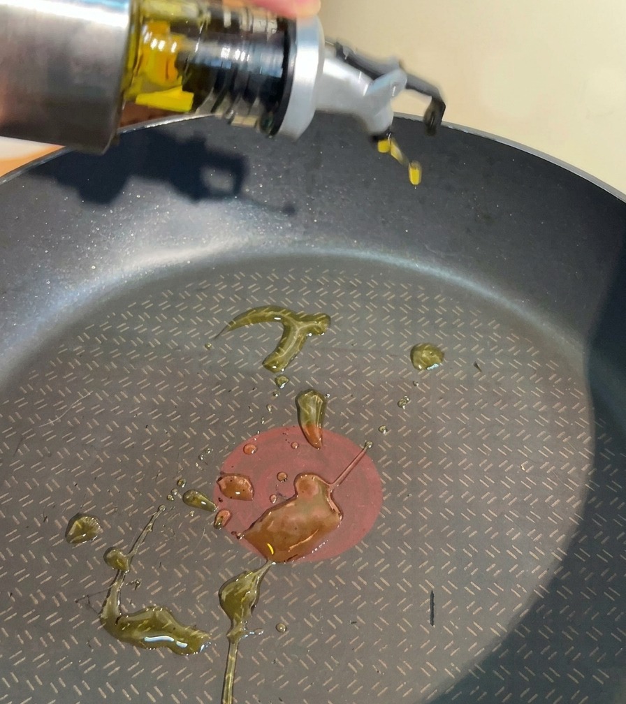 Une main versant de lhuile de cuisson depuis un récipient en inox dans une poêle antiadhésive noire.