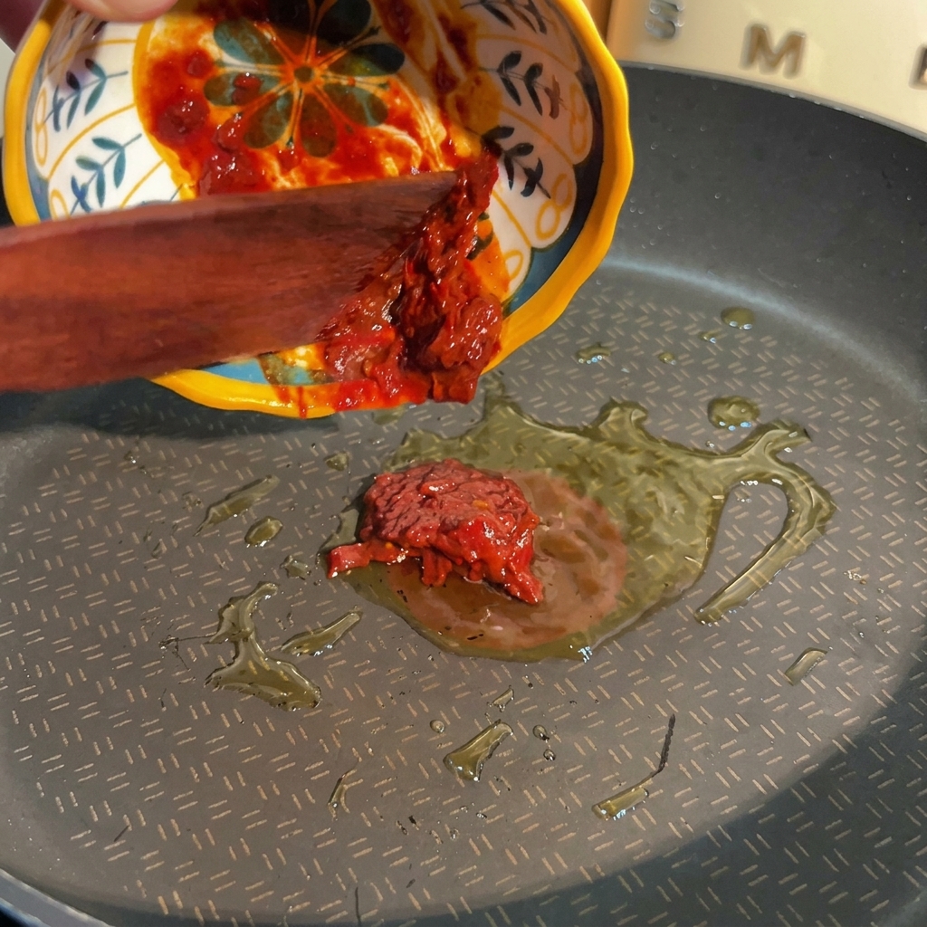 Une spatule en bois poussant une pâte de piment rouge épaisse depuis un bol coloré vers une poêle contenant de lhuile chaude.