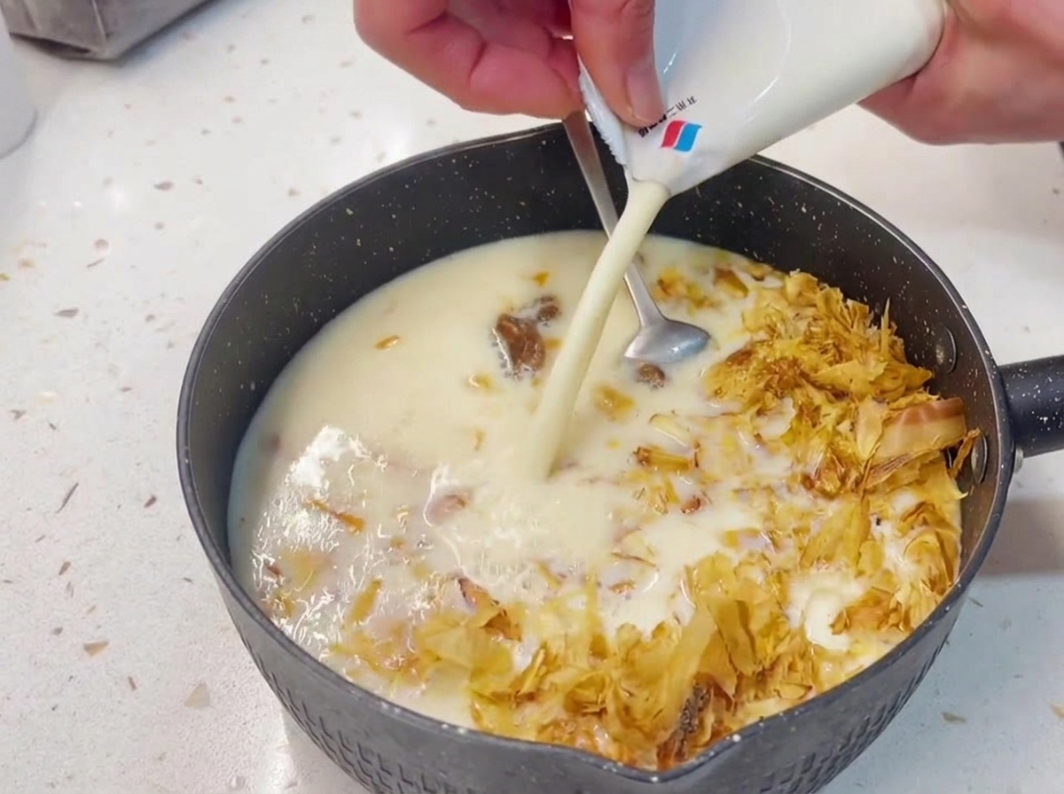 Des mains versant du lait de soja blanc et crémeux depuis sa brique dans une casserole contenant le mélange de thon et de bonite.