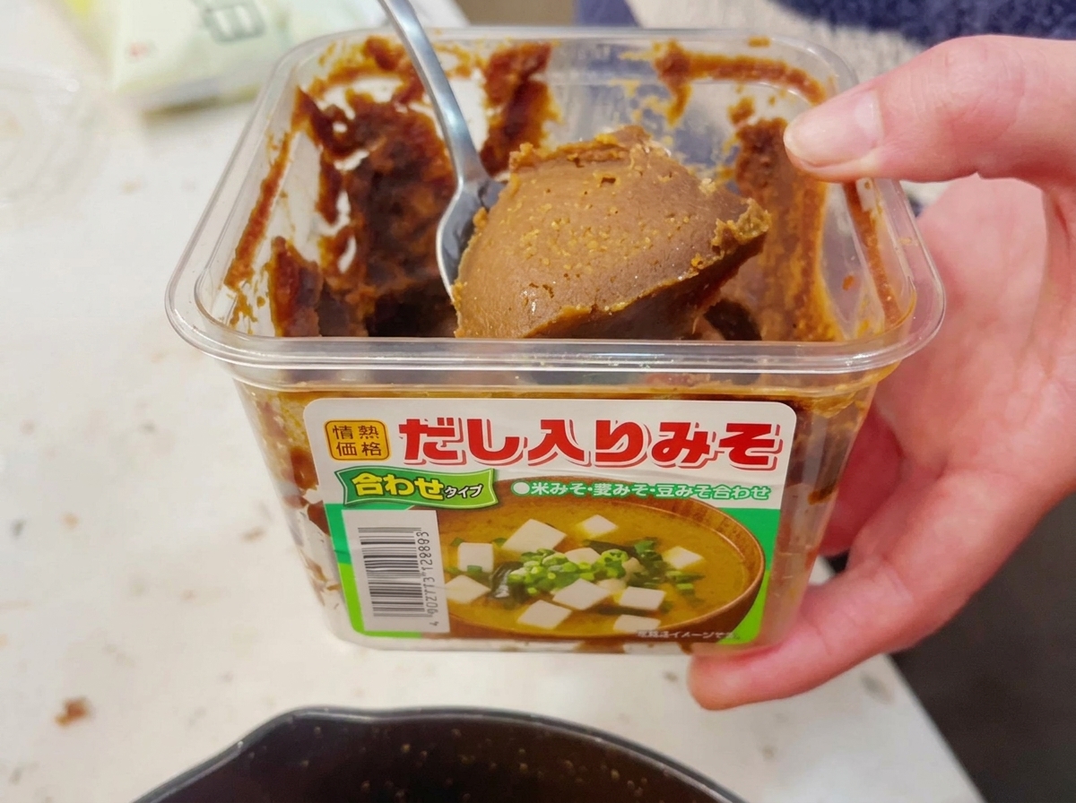 Une main prélevant une généreuse cuillerée de pâte de miso brune et épaisse dans un contenant en plastique transparent avec une cuillère en métal.