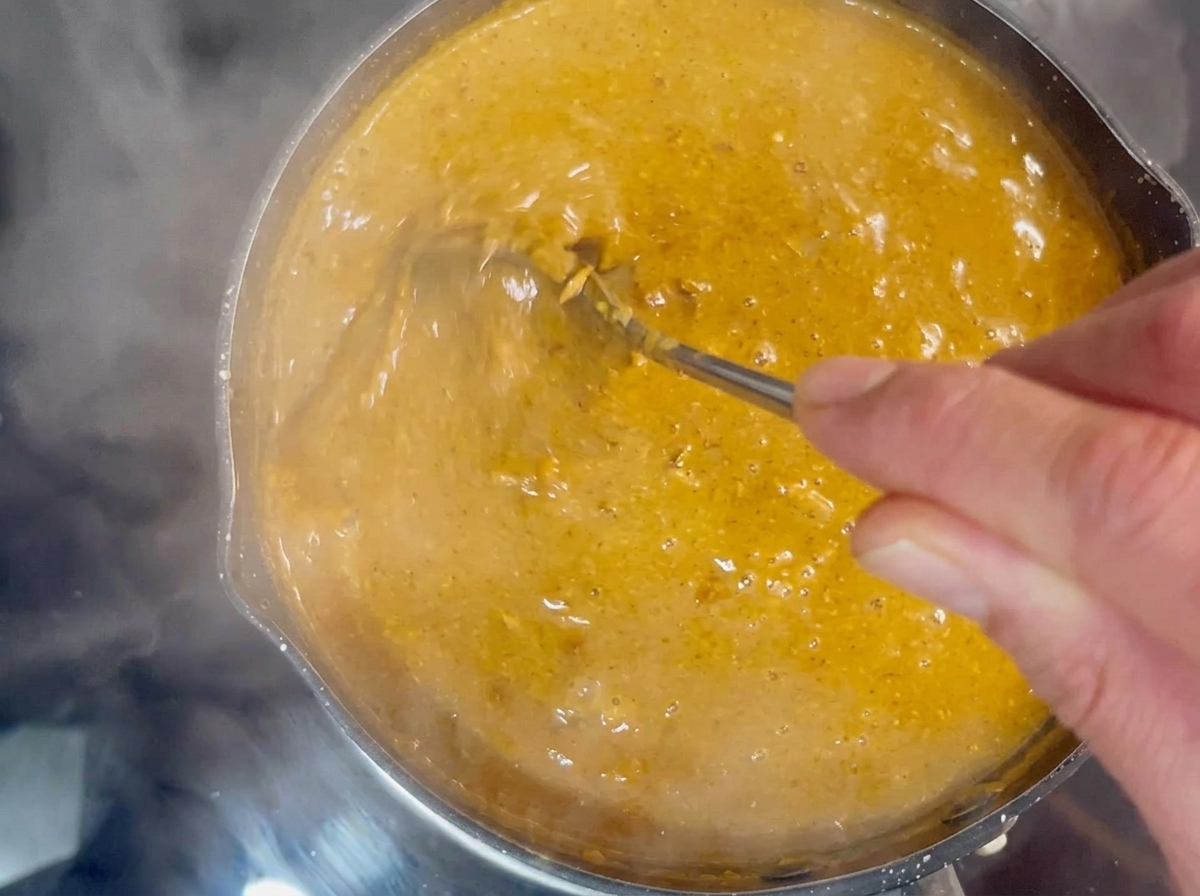 Une main utilisant une cuillère en métal pour remuer un bouillon épais et bouillonnant de couleur orange-brun dans une petite casserole.
