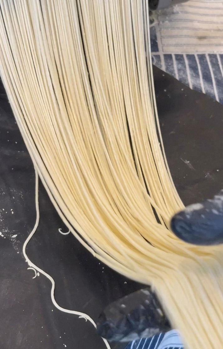 De longs brins de nouilles de ramen jaune pâle sortant dune machine en métal sur une surface sombre.