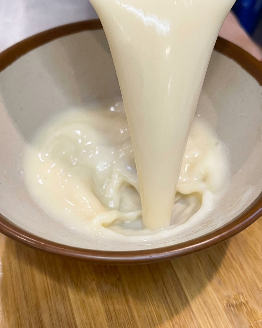 Un filet continu de bouillon de soupe crémeux et pâle versé dans un bol en céramique aux bords marrons.