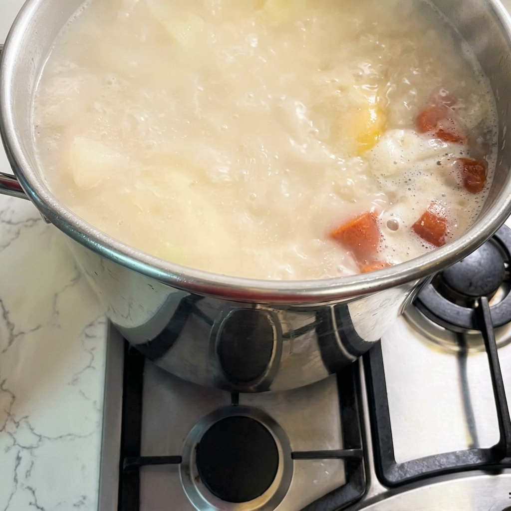 Une grande marmite en acier inoxydable sur une gazinière remplie dun bouillon frémissant, de couleur blanc crème et orangé.
