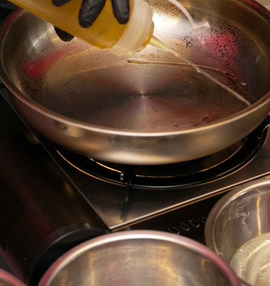 De lhuile de cuisson claire versée depuis un flacon souple en plastique dans une poêle étincelante en acier inoxydable sur un réchaud portable.