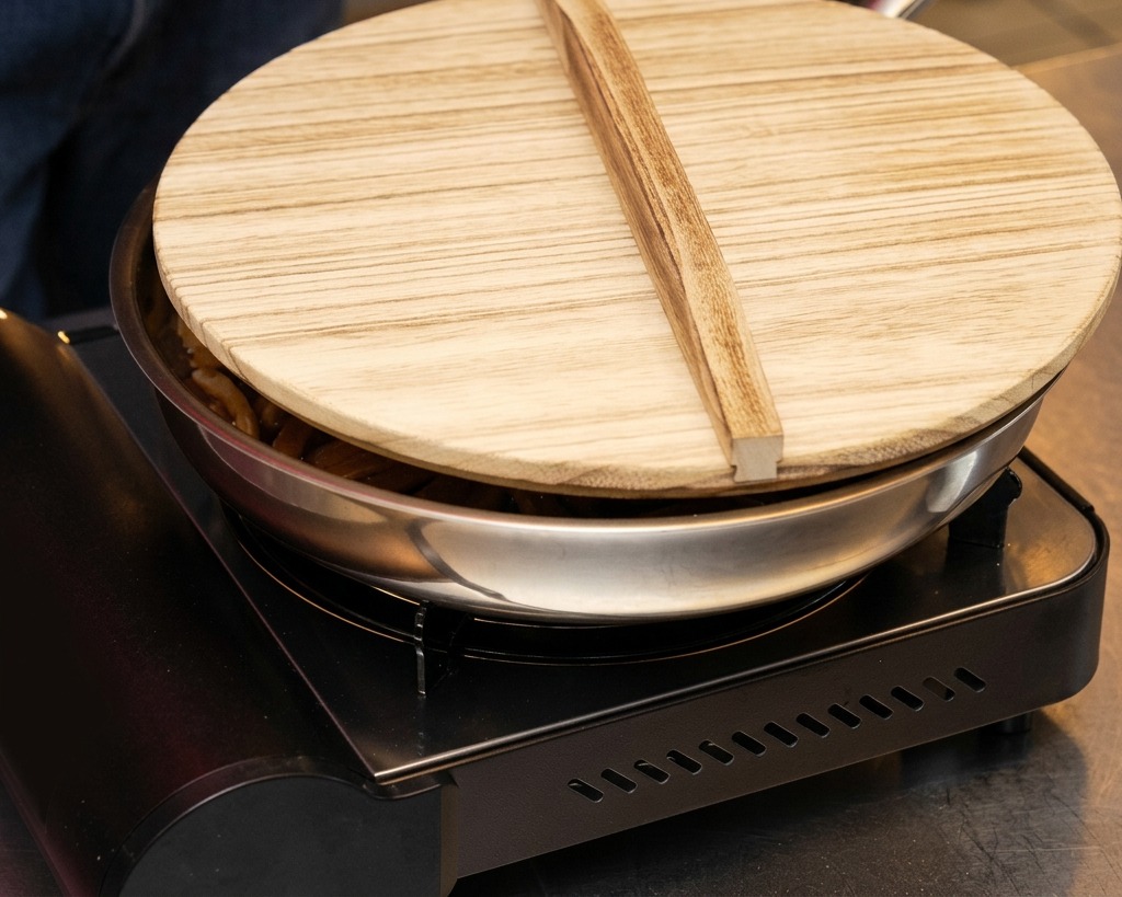 Une poêle en acier inoxydable contenant les pousses de bambou sur un réchaud à gaz portable, couverte par un couvercle en bois traditionnel.