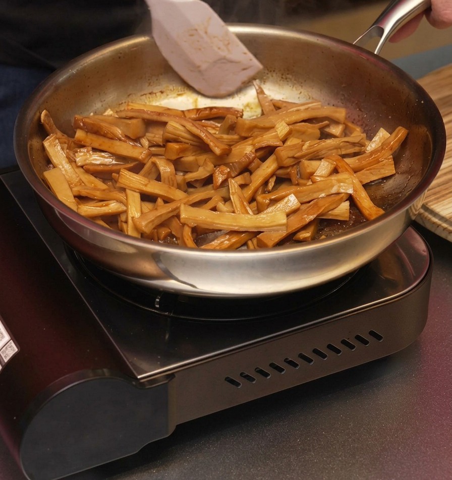 Une spatule blanche mélangeant les pousses de bambou brunes et glacées dans une poêle pour sassurer quelles sont uniformément enrobées dhuile de sésame aromatique.