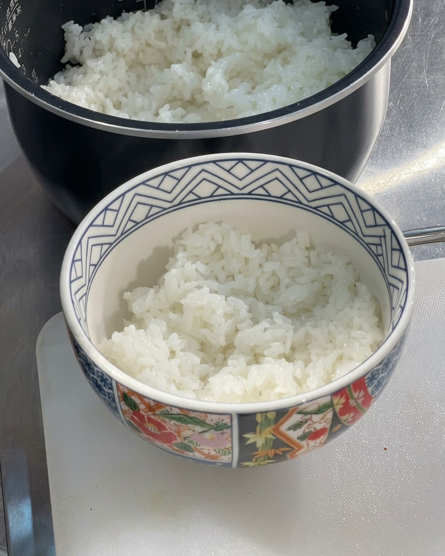 Un bol en céramique à motifs à moitié rempli de riz blanc chaud et moelleux, posé sur une planche à découper à côté dun cuiseur à riz.
