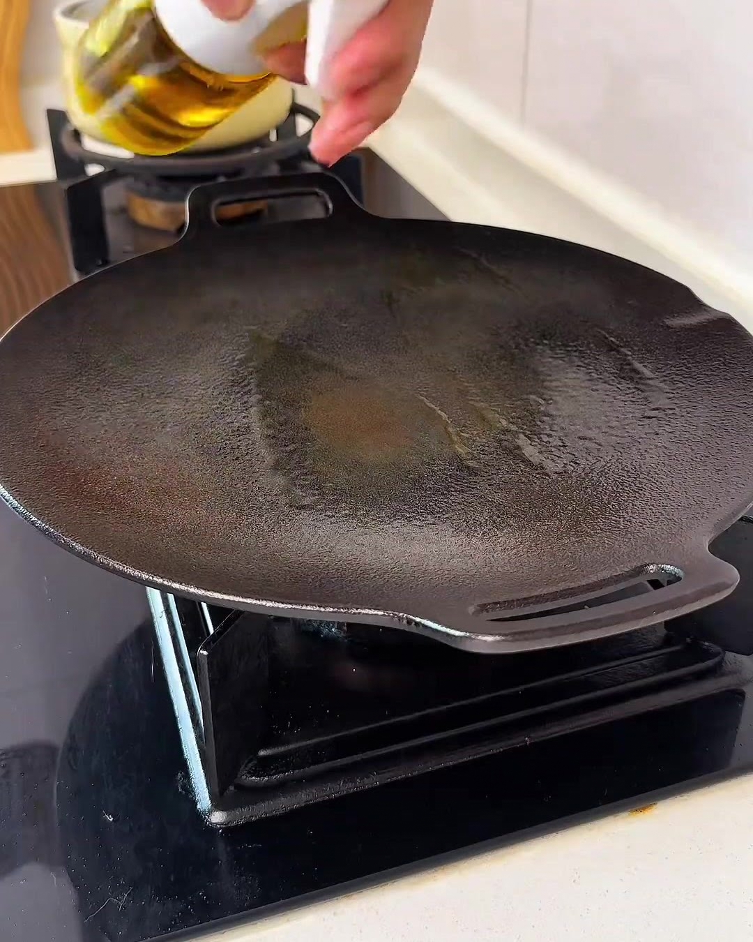 Une main vaporisant une fine brume dhuile de cuisson dorée sur une poêle en fonte noire propre.