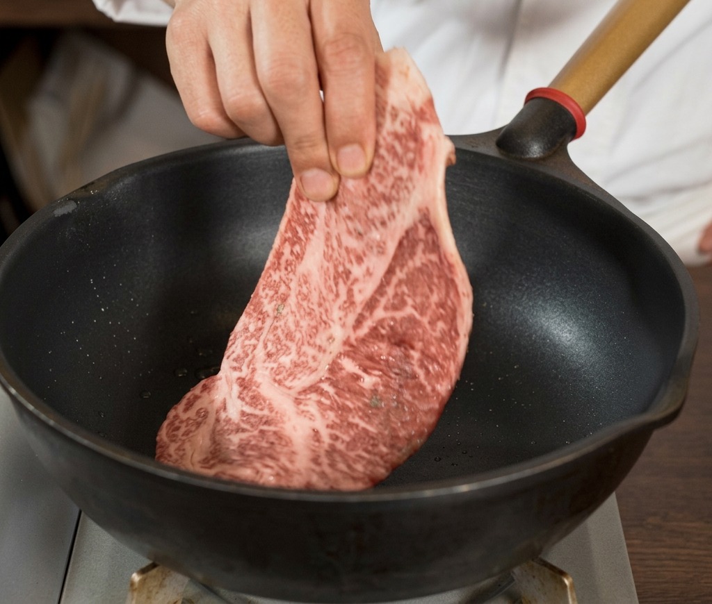 Une main déposant un grand steak de bœuf cru bien persillé et assaisonné de poivre noir dans une poêle noire bien chaude.