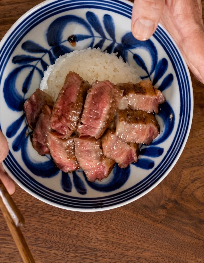 Vue de dessus dune main tenant un bol aux motifs bleus alors que des tranches de steak sont disposées en éventail sur un lit de riz blanc.