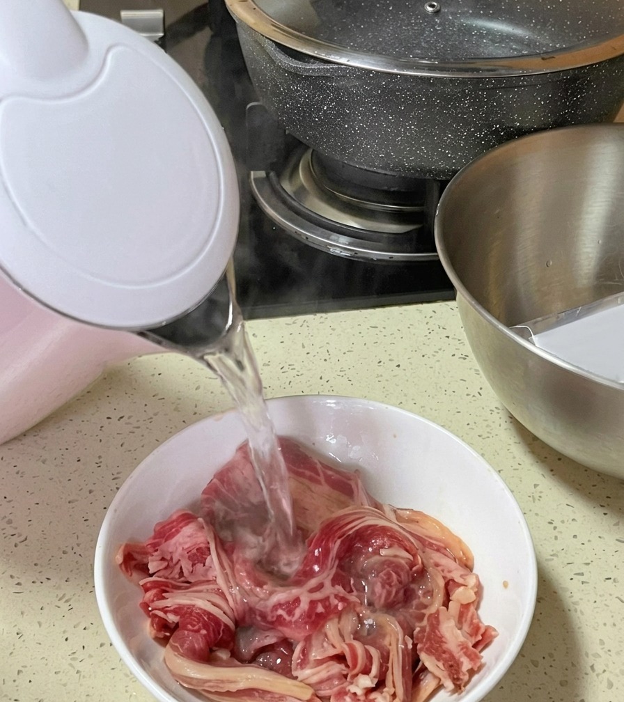 De leau bouillante fumante versée depuis une bouilloire électrique rose et blanche sur de fines lamelles de bœuf cru dans un bol.