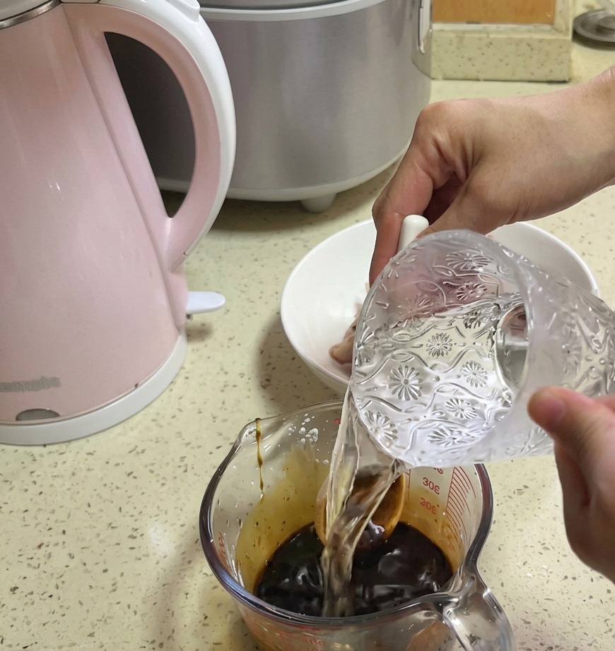 De leau claire versée depuis une carafe en verre décorée dans un verre mesureur contenant une base de sauce marron foncé.