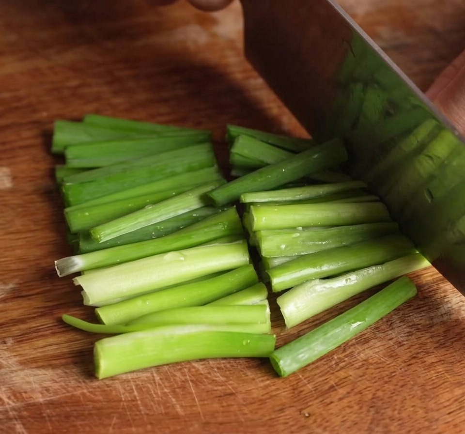 Un couteau coupant une botte de ciboulette vert vif en courts tronçons sur une planche en bois.