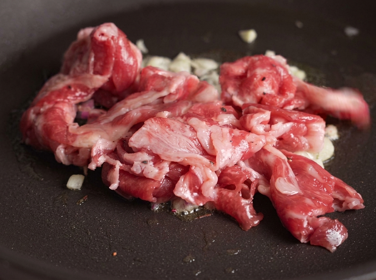 Tranches de bœuf cru ajoutées dans une poêle contenant de lail doré, la viande commençant à brunir sous leffet de la chaleur.