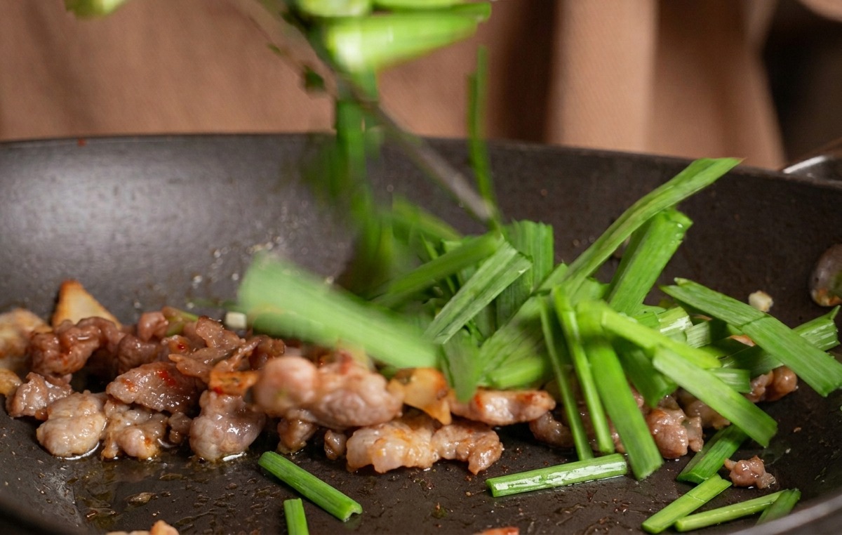 Une main parsemant des morceaux de ciboulette verte dans une poêle contenant un sauté de bœuf et de kimchi.