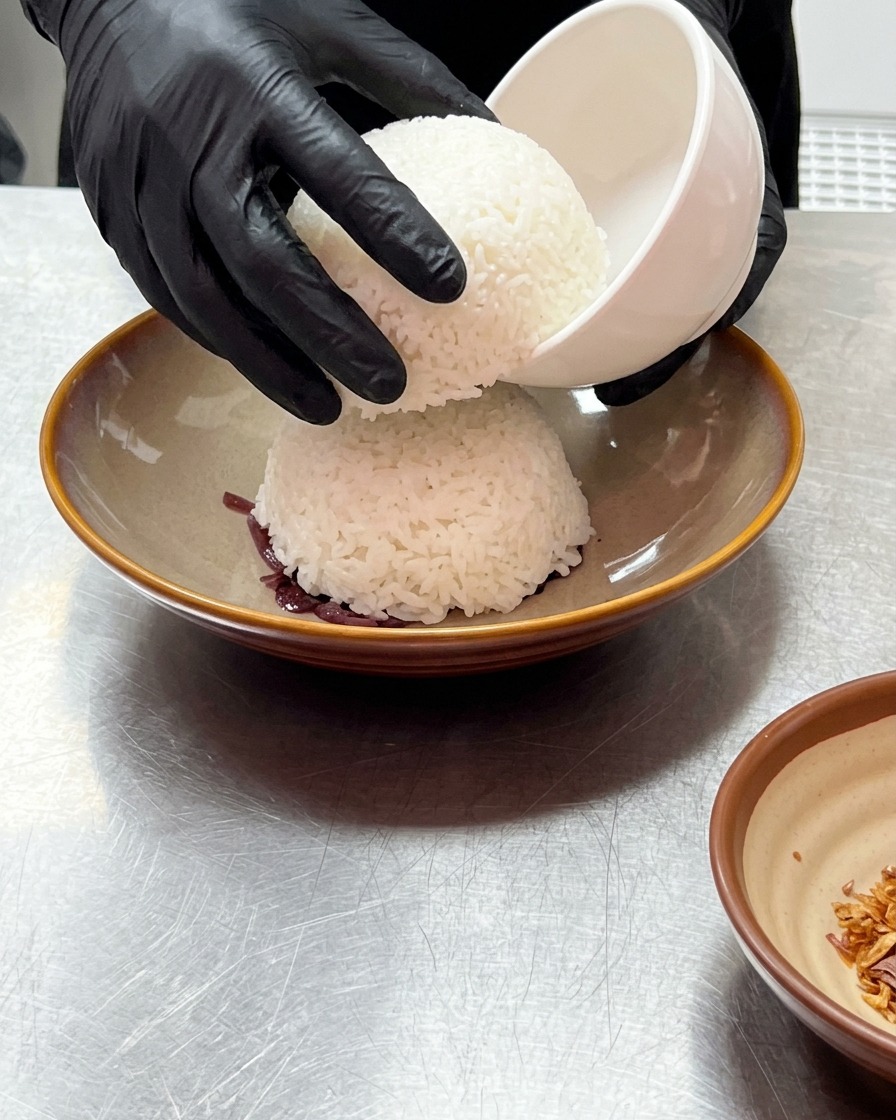 Des mains gantées soulevant un petit bol blanc pour révéler un dôme de riz blanc cuit à la vapeur posé sur les oignons rouges.