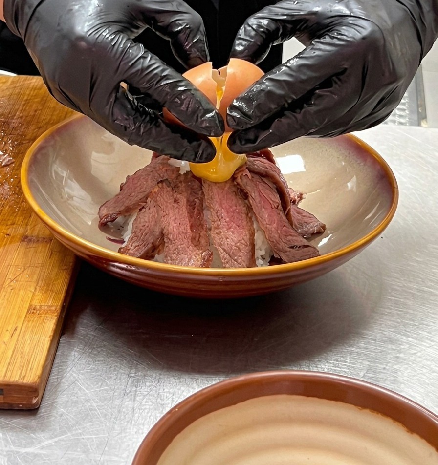 Deux mains gantées cassant délicatement un œuf onsen partiellement cuit au sommet dune montagne de lamelles de bœuf.
