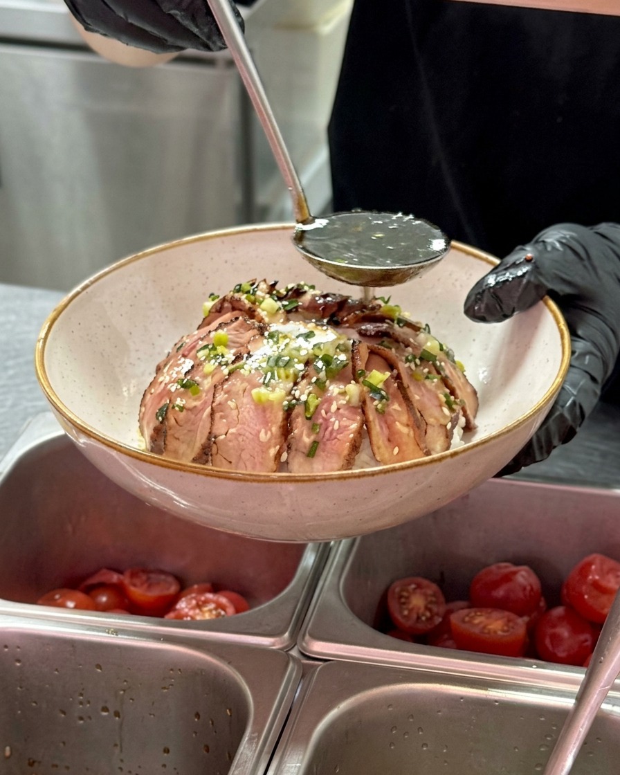 Une louche en métal versant une sauce sombre et brillante sur un bol de bœuf émincé disposé autour dun œuf, avec des morceaux de cébette visibles.