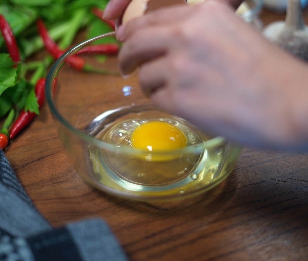 Mains cassant délicatement une coquille dœuf brun au-dessus dun bol en verre transparent contenant déjà un œuf entier.