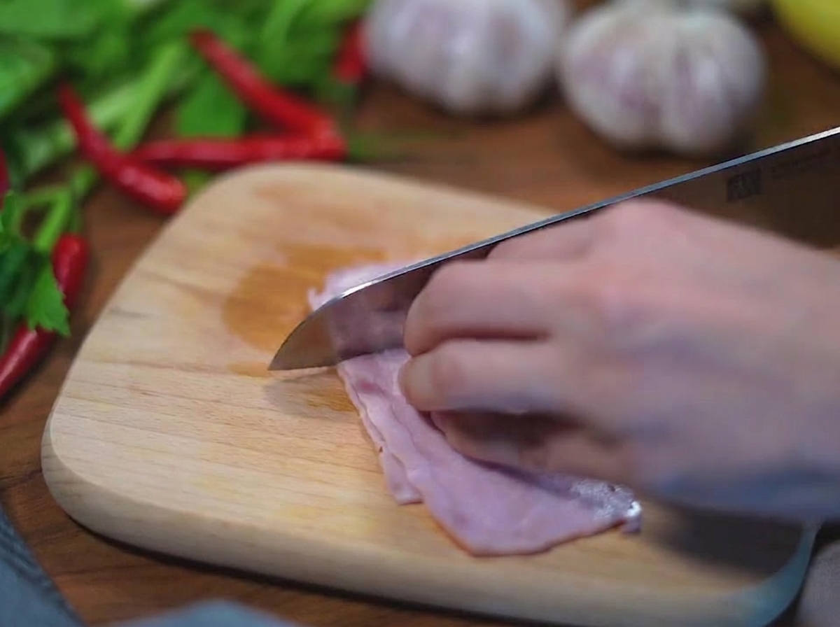 Gros plan sur des mains coupant des tranches de bacon cru en petits carrés réguliers avec un couteau en acier inoxydable sur une planche en bois.