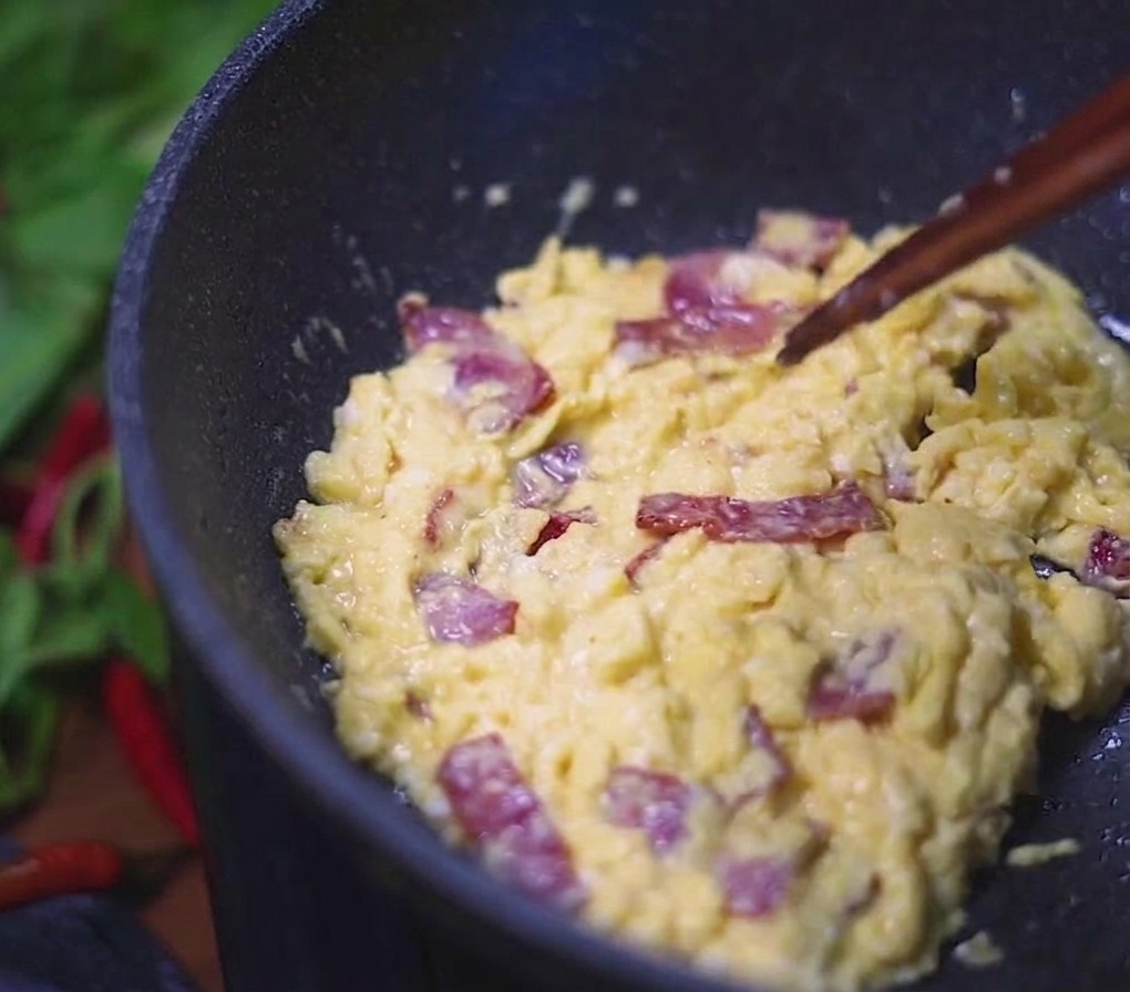 Œufs brouillés crémeux mélangés à des morceaux de bacon savoureux, remués à laide de baguettes dans une poêle sombre.