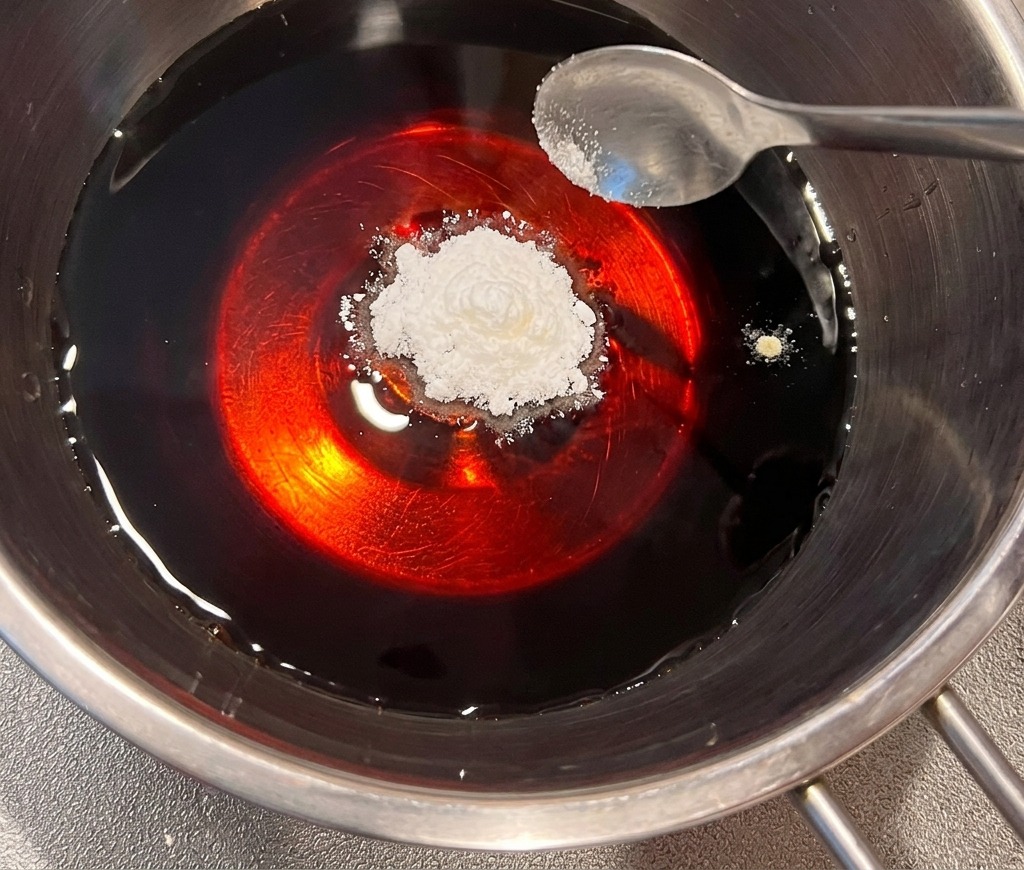 Une cuillerée de dashi en poudre ajoutée à un liquide sombre à base de soja dans une casserole en acier inoxydable.