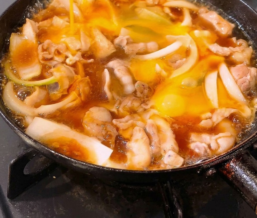Œufs battus versés dans une poêle remplie de poitrine de porc et doignons qui mijotent dans une sauce au dashi.