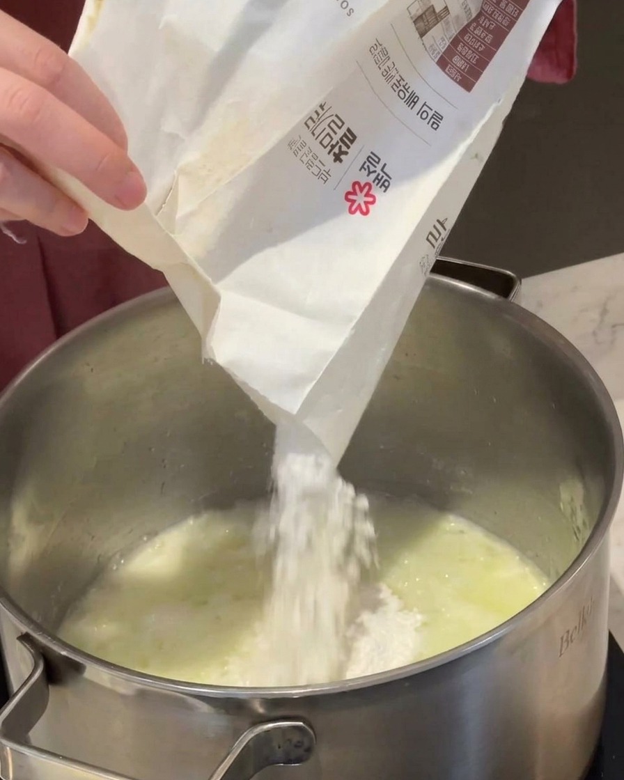 De la farine blanche versée depuis un sachet en papier dans une casserole contenant du beurre fondu et des oignons.