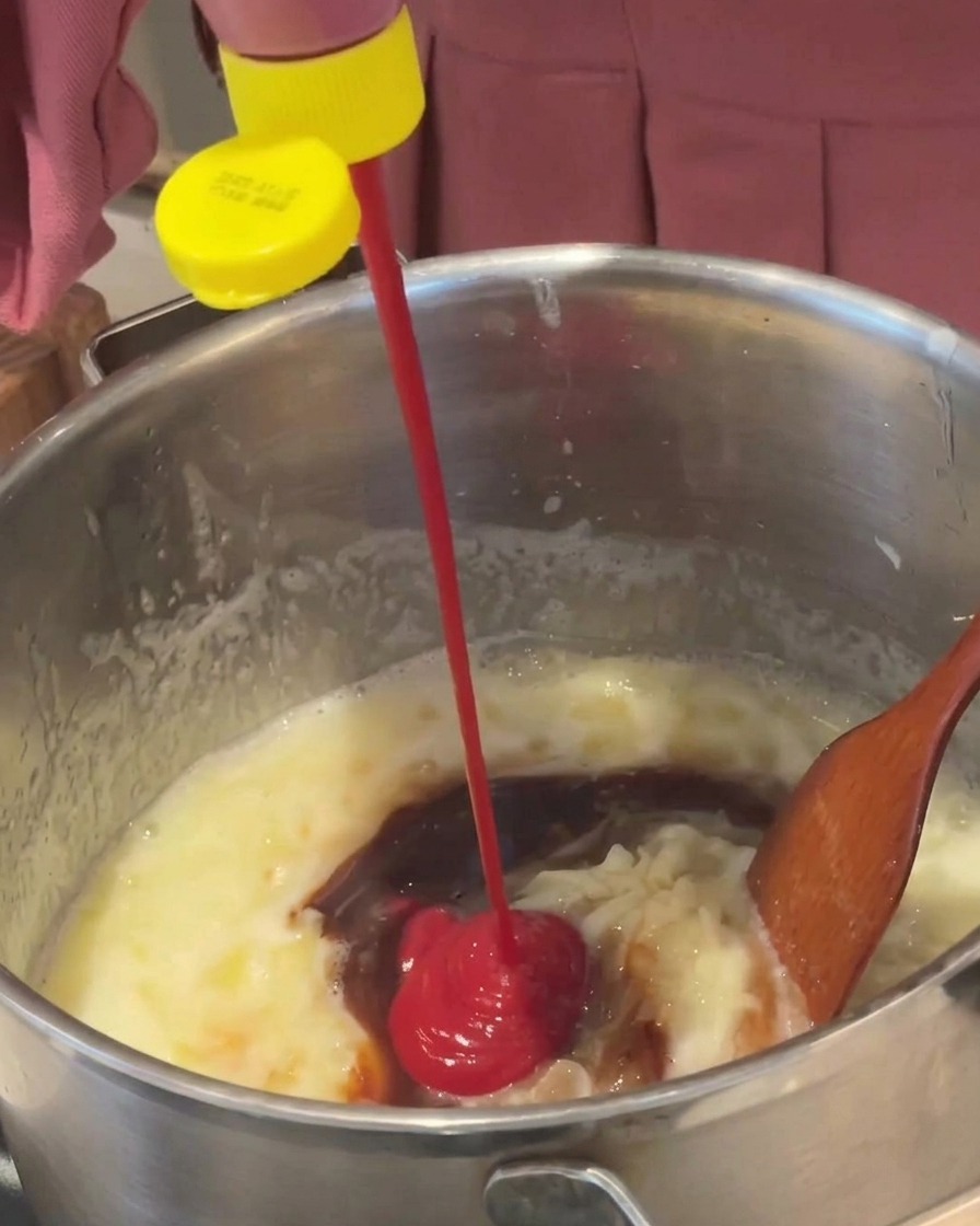 Du ketchup rouge vif pressé depuis une bouteille au bouchon jaune dans une sauce brune qui mijote dans une casserole.