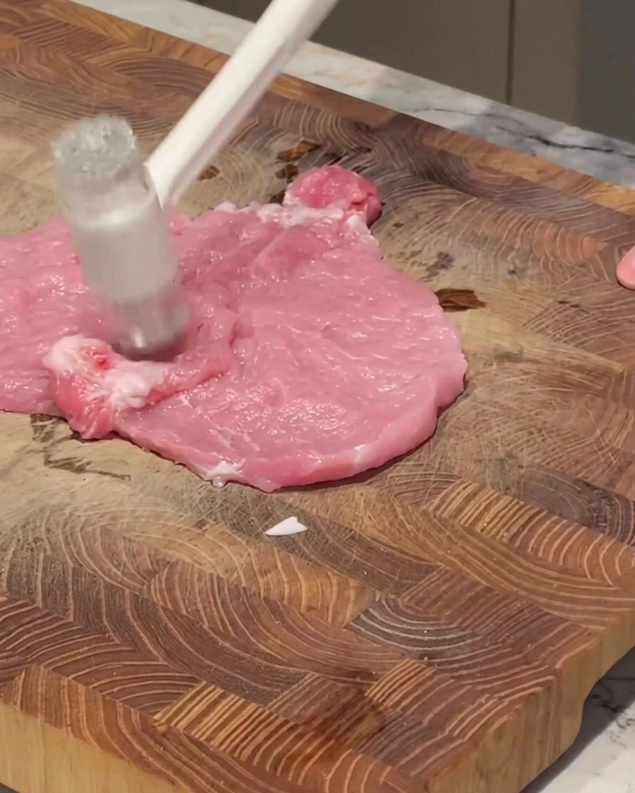 Un attendrisseur à viande blanc frappant un morceau de porc rose cru sur une planche à découper en bois.
