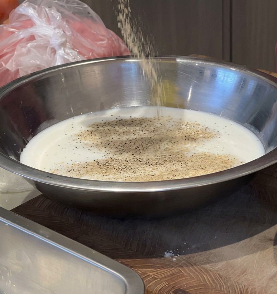 Une personne versant des assaisonnements depuis un récipient dans un bol en inox rempli de lait pour créer une marinade.